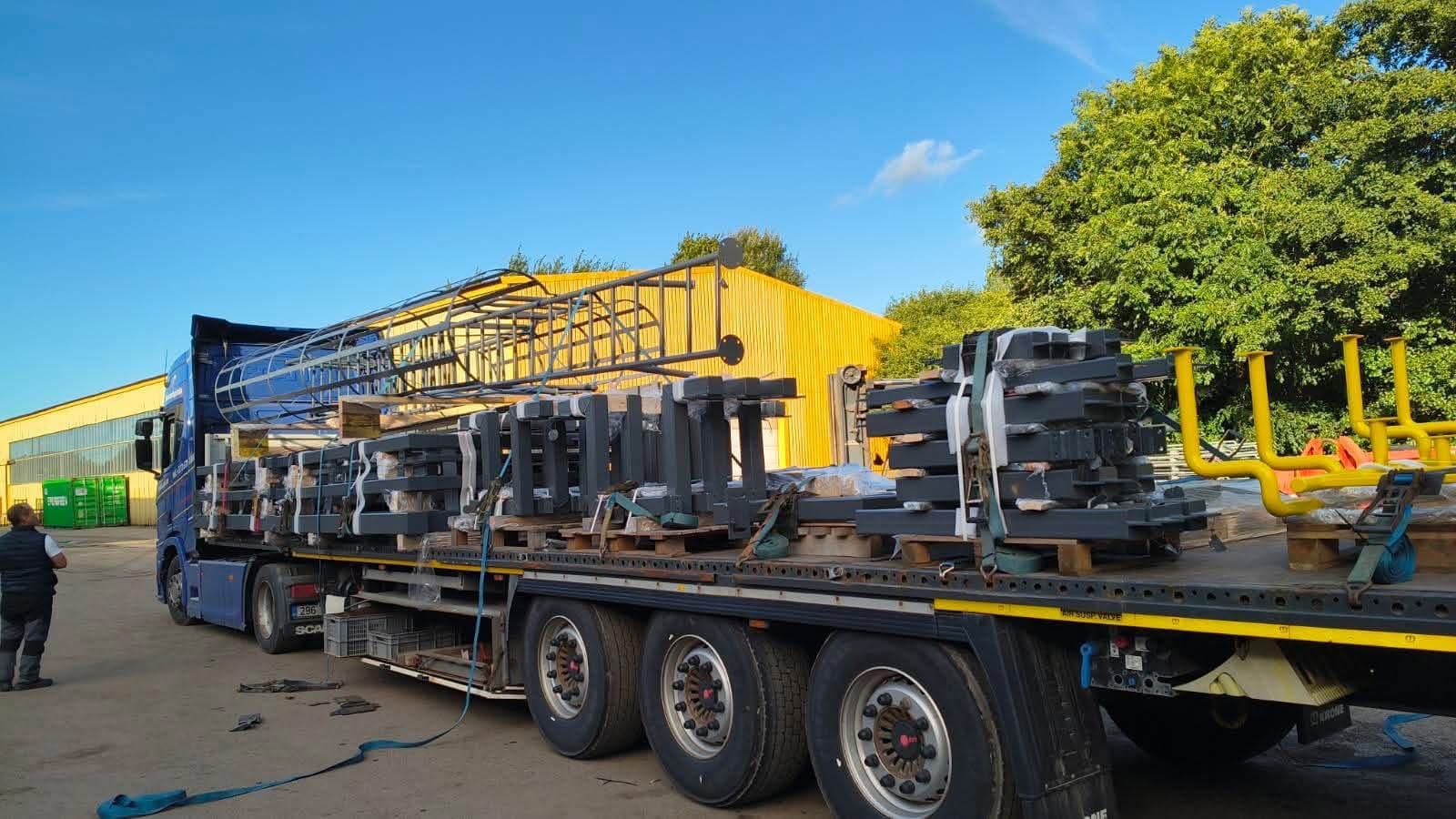 Flatbed trailer loaded with dark steel structure parts and yellow frames at the Novatex yard in Tallinn